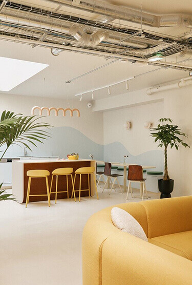 Cosy office canteen with yellow sofa, yellow bar stools, wooden chairs and plants.