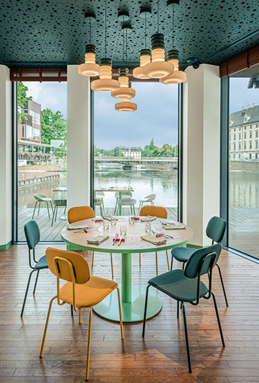 A cosy restaurant overlooking the river, furnished with yellow and green chairs and a round table, lit by designer ceiling lamps.