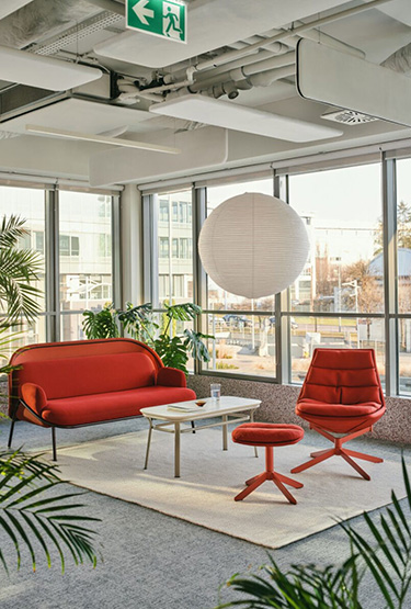 A bright relaxation area with a red armchair and a red sofa, surrounded by plants, with large glass windows and a paper ceiling lamp.