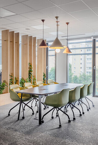 A modern meeting room with a large table, green chairs on wheels and hanging lamps.