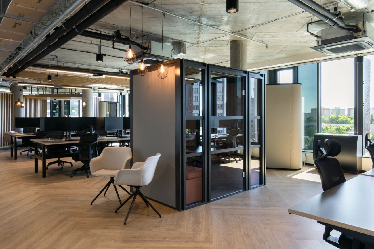 Biuro typu open-space wyposażone w biurka z panelami oraz szarą, przeszkloną budkę akustyczną