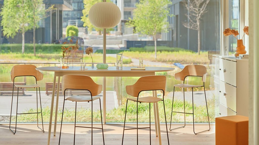 Holzbarhocker Afi mit Rückenlehne an einem hohen Tisch in einem hellen Innenraum mit großen Fenstern.