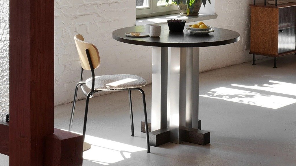 A round table with a wooden top and a metal base, paired with a chair in a bright interior